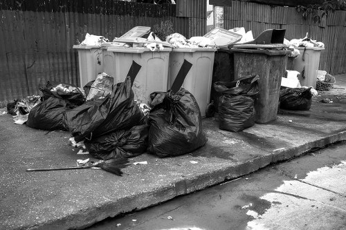 A collection of large black and grey rubbish bags filled with waste are piled on a paved outdoor area, with some bags leaning against several large wheelie bins and a smaller bin. The bins are situated on a concrete driveway or pathway, with a wooden fence in the background. The grey bins have lids open or partially open, revealing scattered paper, cardboard, and miscellaneous rubbish on top or inside them. The black bags appear to contain mixed waste, some of which has spilled onto the ground nearby. The overall scene suggests a recent rubbish removal effort, possibly part of a garden or property clearance service in Lewisham, with the environment indicated by residential fencing and an outdoor setting suitable for waste collection.