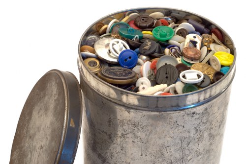 A round, metal rubbish bin filled to the brim with a variety of plastic buttons of different sizes, shapes, and colours, including blue, black, white, green, and brown. The surface of the bin shows signs of wear with scratches and a slightly tarnished appearance. The bin is positioned outdoors on a paved area, with the lid resting against its side, and the background is plain white. This image relates to waste collection and disposal services, such as those offered by Garden Clearance Lewisham, often involving the removal of miscellaneous household or garden waste materials in Lewisham or surrounding areas within the London borough.