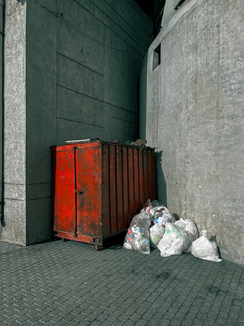 Three large black and white plastic trash bags filled with waste are placed on the pavement next to a curb, partially leaning against a black metal fence with vertical bars. One bag appears to be tied