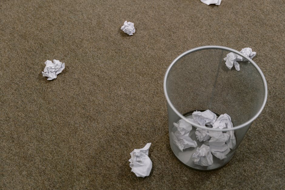 A clear, cylindrical trash bin made of lightweight plastic with a smooth, semi-transparent surface and a rounded rim, positioned on a brown textured carpeted floor. Inside the bin is crumpled white pa